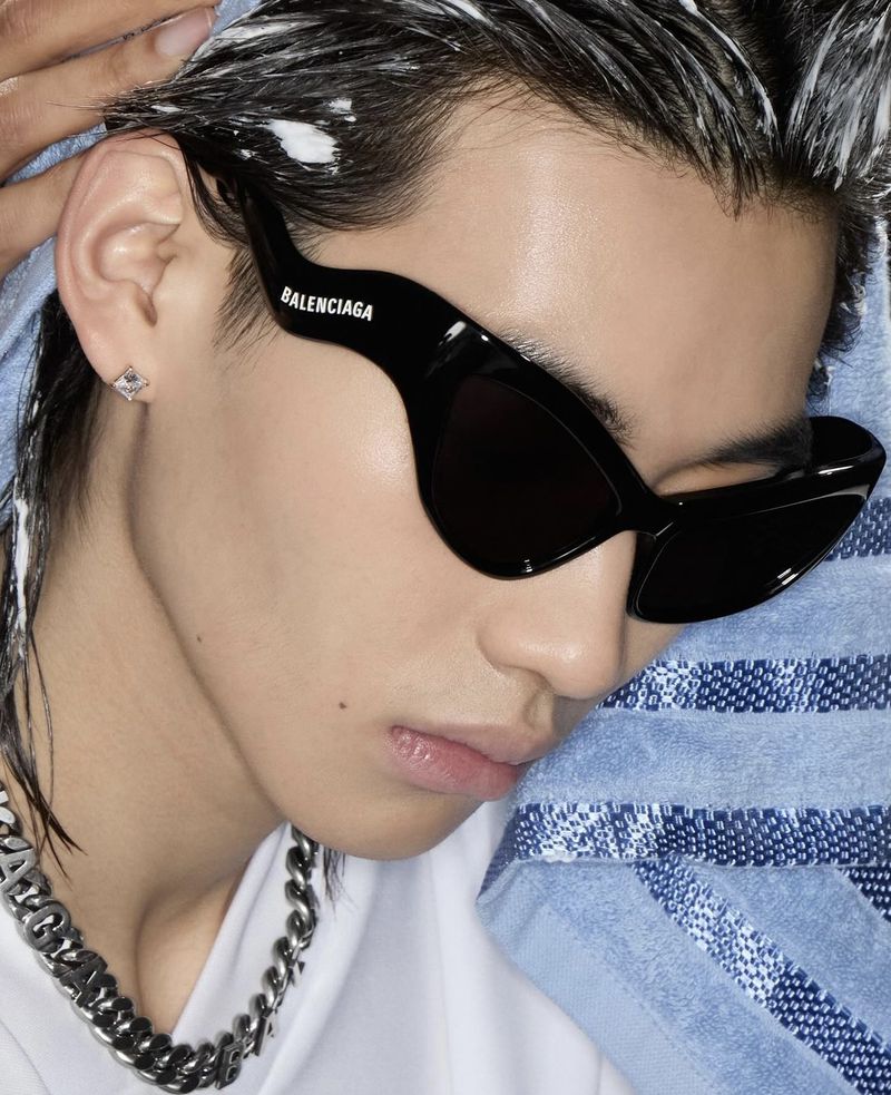 Close-up of a person wearing Balenciaga sunglasses and a silver chain necklace, with a blue towel and light-colored clothing.