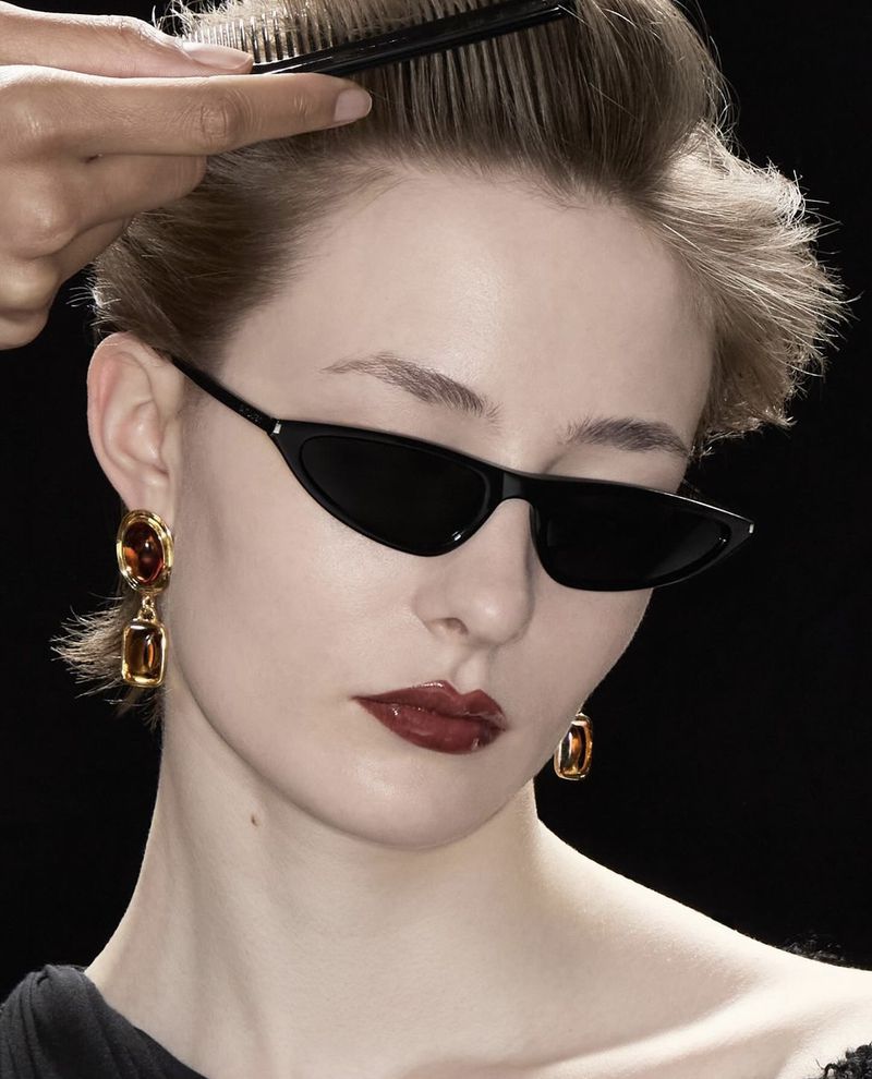 Close-up of a woman with short hair and black sunglasses getting her hair combed in a studio with a black background.