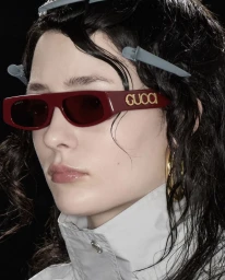 Close-up of model wearing burgundy Gucci rectangular sunglasses with gold branding and grey hair clips against a black background.