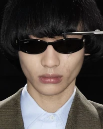 Close-up portrait of an East Asian model wearing black oval sunglasses getting bangs trimmed with scissors against a black background.