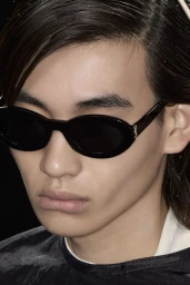 Close-up of a model wearing black oval sunglasses with gold temple detailing against a dark studio background.