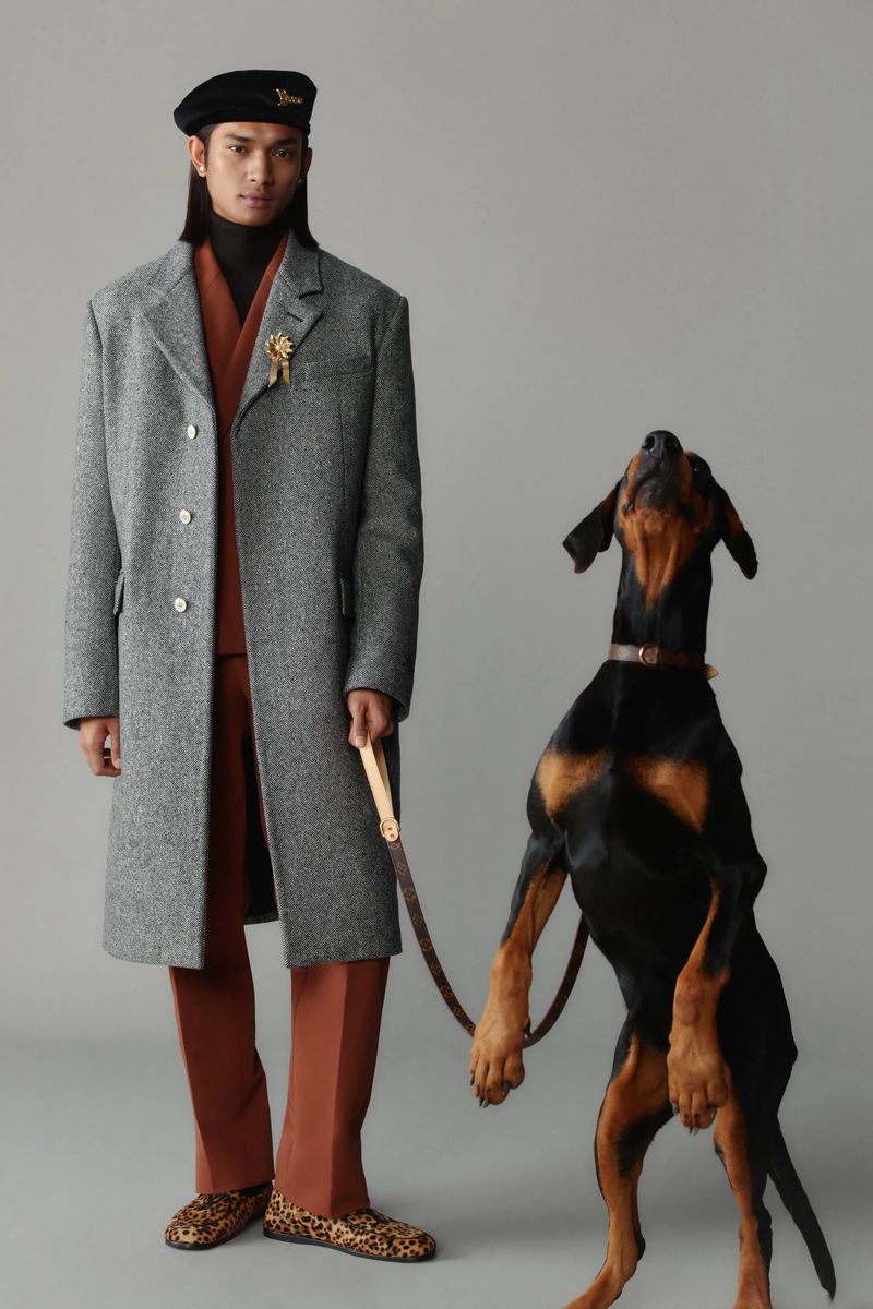 A stylish man poses next to a sleek Doberman dog, both in a fashion forward studio portrait with cool grey tones and modern aesthetic.