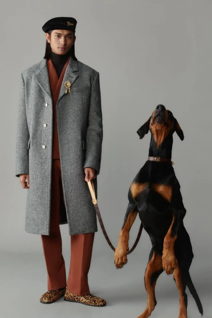 A stylish man poses next to a sleek Doberman dog, both in a fashion forward studio portrait with cool grey tones and modern aesthetic.
