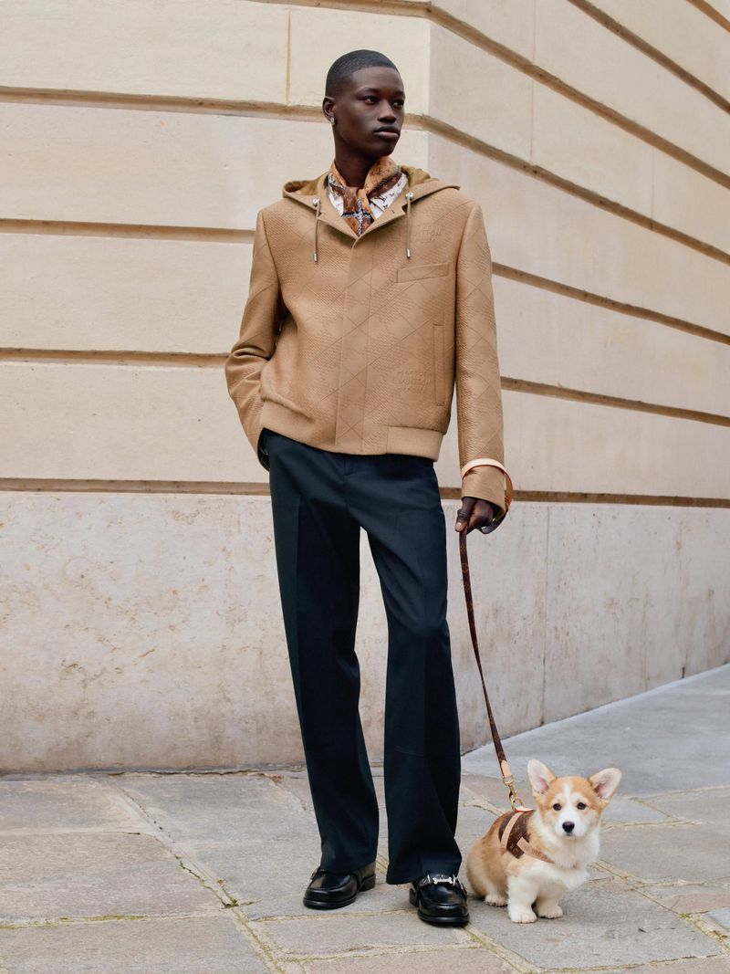 Adult male in hooded jacket and dress pants walks a corgi on a leash along a building in an outdoor urban setting on a sunny day.