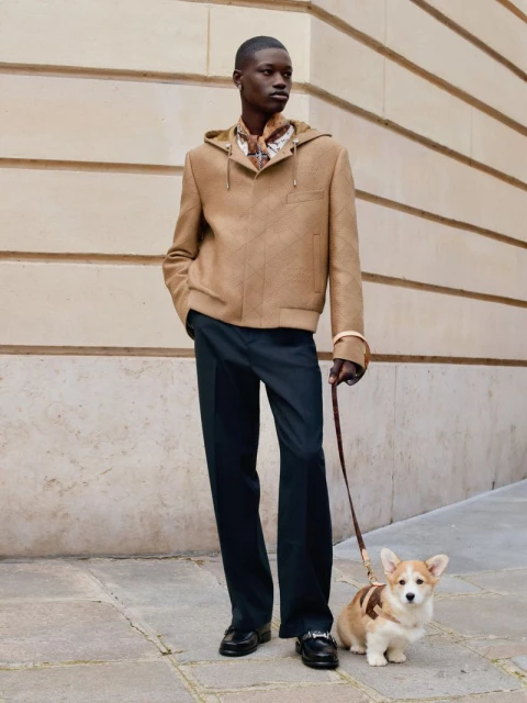 Adult male in hooded jacket and dress pants walks a corgi on a leash along a building in an outdoor urban setting on a sunny day.