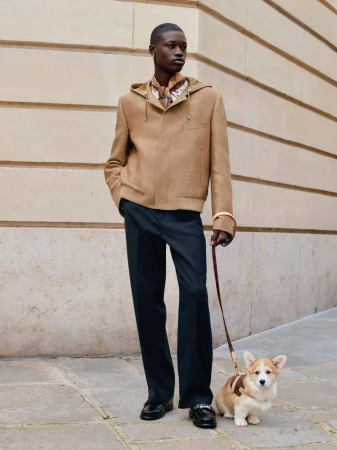 Adult male in hooded jacket and dress pants walks a corgi on a leash along a building in an outdoor urban setting on a sunny day.