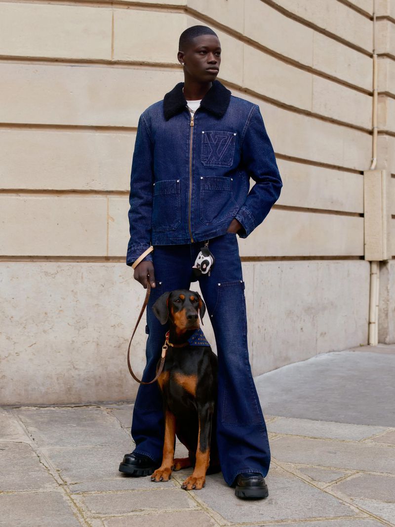 Male model in blue denim jacket and pants poses with a black and tan dog on a city street in natural light.