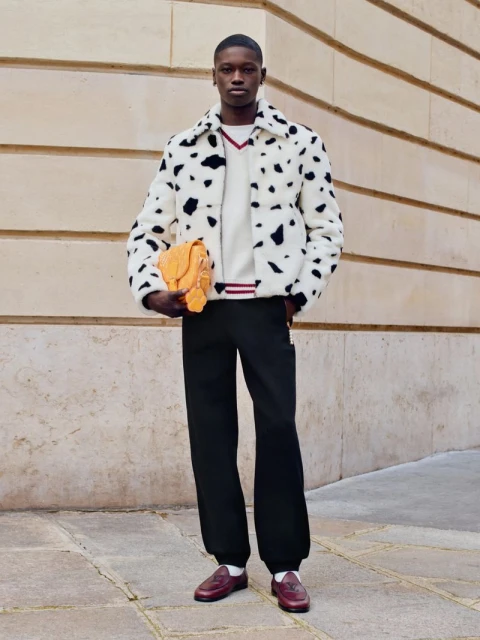 Stylish adult man in a dalmatian print jacket holds a yellow handbag outdoors, complemented by black trousers and brown loafers.