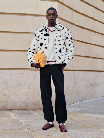 Stylish adult man in a dalmatian print jacket holds a yellow handbag outdoors, complemented by black trousers and brown loafers.