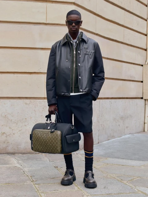 Dark-skinned male model in a black leather jacket, shorts, and sunglasses holding a Louis Vuitton travel bag, posing outdoors.