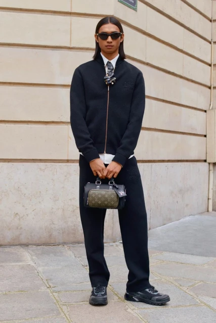 Adult male wearing a black bomber jacket and sweatpants with sunglasses, holding a small handbag, standing in an urban environment.