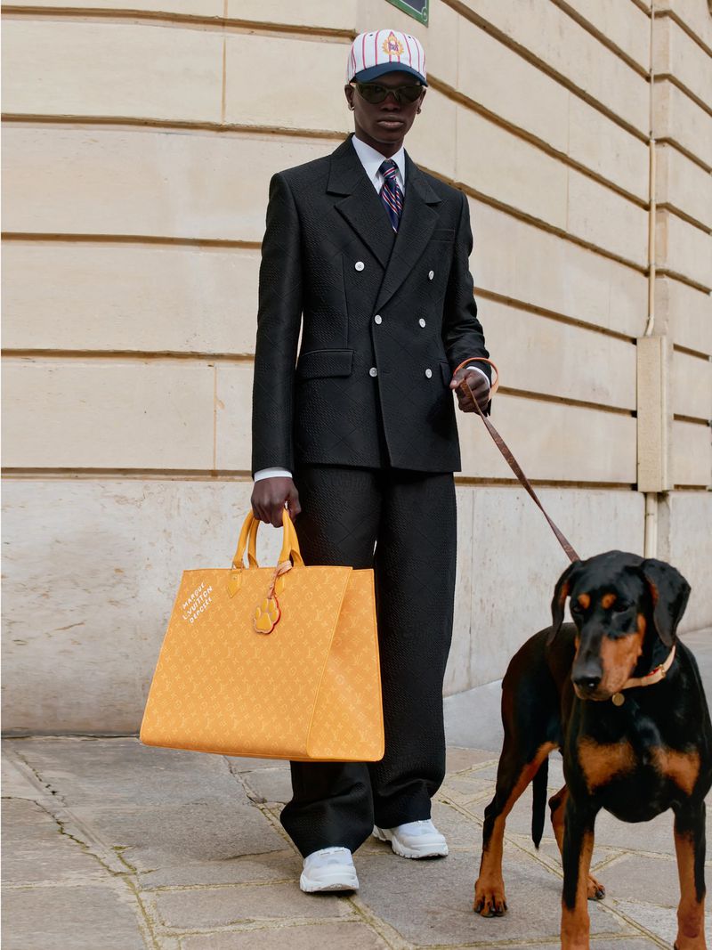 Elegant man in a suit walks a Doberman dog while carrying a large yellow tote bag on a city street with classic architecture.