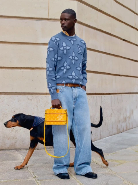 An African-American man in designer jeans and sweater poses outdoors with a Doberman Pinscher dog and a yellow box-shaped handbag.