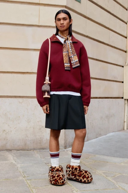 Male fashion model poses in a burgundy sweater, black shorts, socks, and leopard-print shoes in an urban setting with a purse.