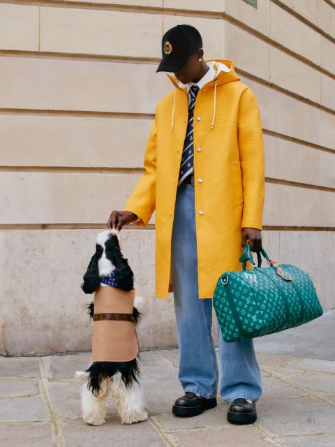 Man in a yellow coat pets a black and white dog with a tan coat in front of a neutral wall with a green duffel bag.