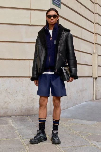 Male fashion model wearing a black shearling jacket, navy shorts, and black sneakers stands in front of a stone building.