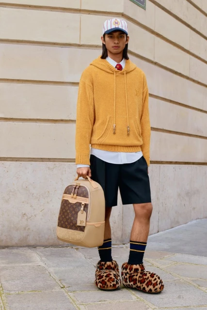 Full shot of a young adult male in casual fashion, wearing a baseball cap, hooded sweatshirt, shorts, socks, and slippers, holding a tan backpack.