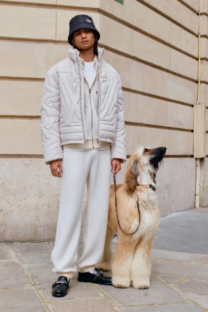 Adult male model poses outdoors with tan Afghan Hound. He wears a quilted jacket, knit bucket hat, and white sweatpants. 