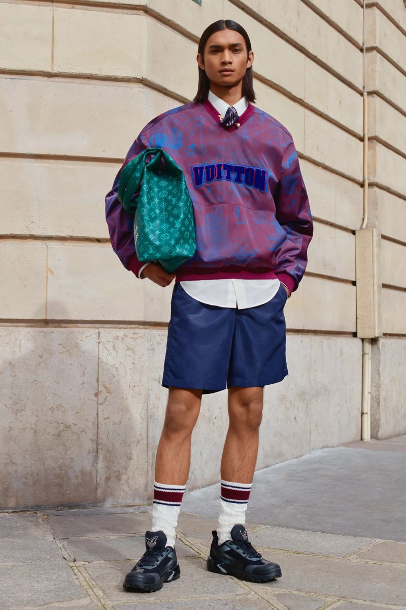 Adult male model wearing a colorful sweatshirt, shorts, socks, and sneakers, carrying a green tote bag while posing outdoors.