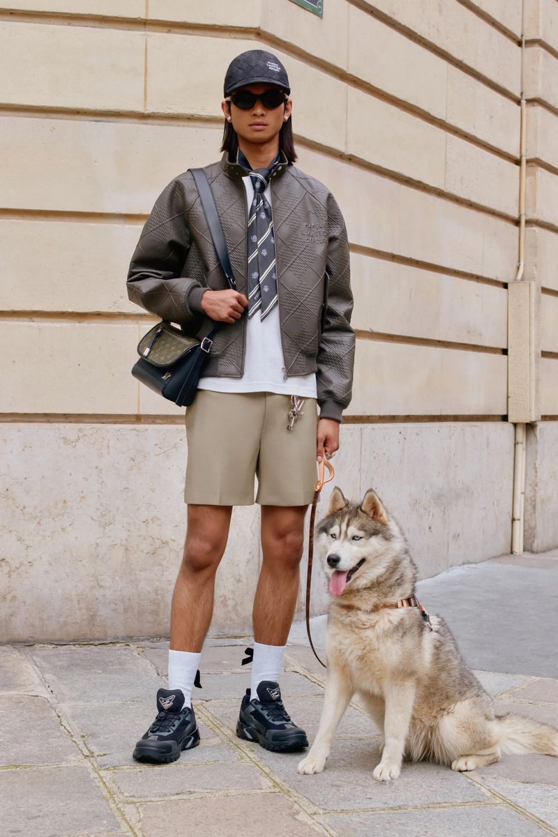 Adult male in a jacket, shorts, sneakers, and baseball cap walks a dog on a city sidewalk next to a tan building.