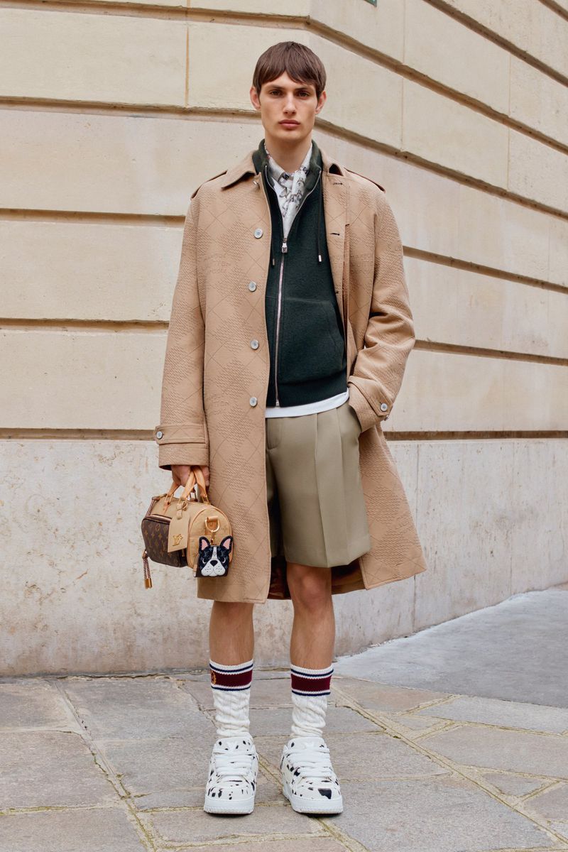 A young adult male stands outdoors wearing a trench coat, shorts, athletic socks and Dalmatian print shoes, holding a handbag.
