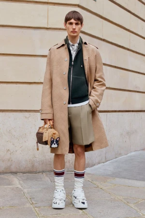 A young adult male stands outdoors wearing a trench coat, shorts, athletic socks and Dalmatian print shoes, holding a handbag.
