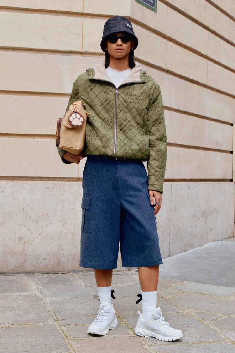 A person standing outdoors on a paved surface, wearing an olive quilted jacket, navy shorts, white socks, and white sneakers.