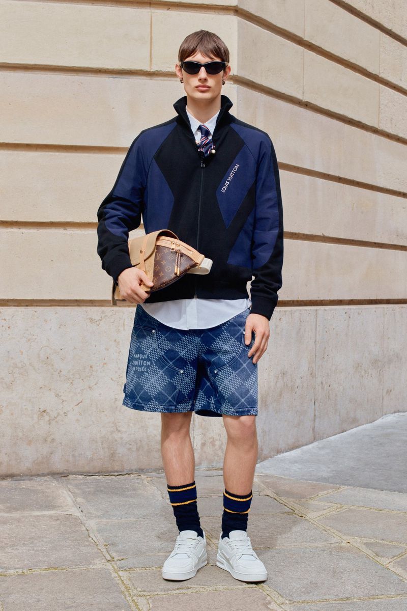 A full shot of an adult male in patterned shorts, a jacket, and white sneakers, holding a bag, posing against a tan building outdoors.