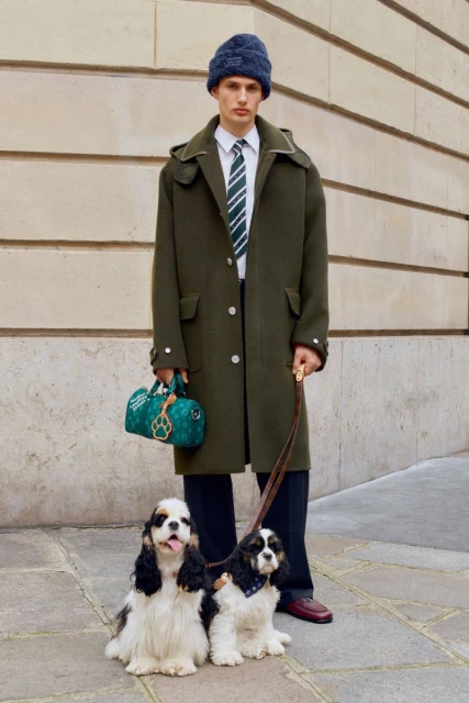 Male fashion model in coat with two dogs on leashes outdoors. He carries a designer bag, wears a striped tie and wool hat.