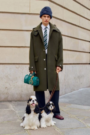 Male fashion model in coat with two dogs on leashes outdoors. He carries a designer bag, wears a striped tie and wool hat.