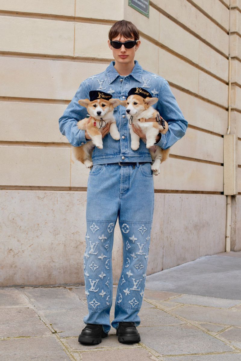 A person in denim outfit stands outdoors holding two small dogs wearing hats, showcasing street fashion with pets and stylish apparel.