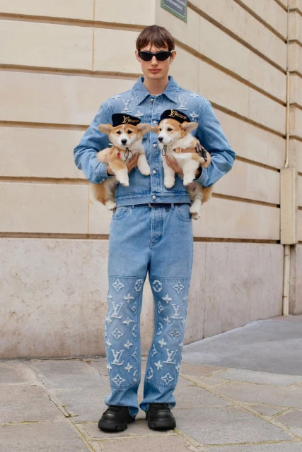 A person in denim outfit stands outdoors holding two small dogs wearing hats, showcasing street fashion with pets and stylish apparel.