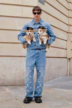 A person in denim outfit stands outdoors holding two small dogs wearing hats, showcasing street fashion with pets and stylish apparel.