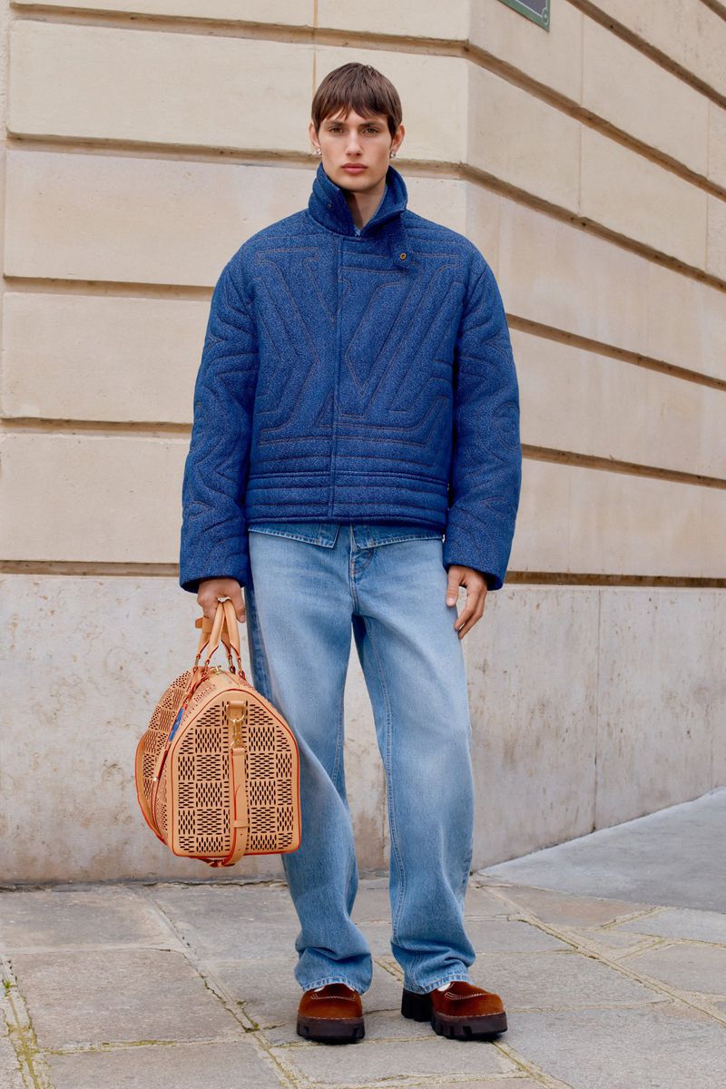 A male model poses in front of a neutral building wearing a blue jacket, loose jeans, and brown loafers, holding a patterned duffel bag.