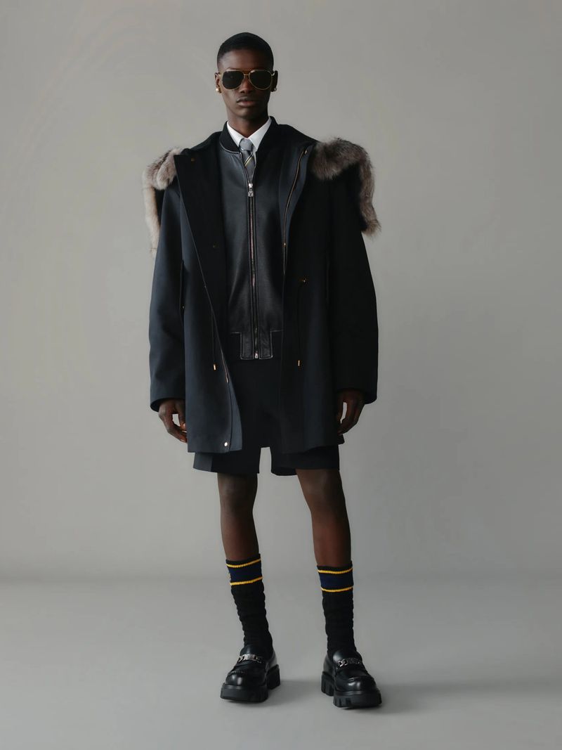 A dark-skinned male model poses in a fur-trimmed coat, shorts, socks, and loafers against a neutral backdrop in a studio setting.