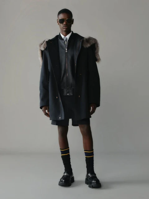 A dark-skinned male model poses in a fur-trimmed coat, shorts, socks, and loafers against a neutral backdrop in a studio setting.
