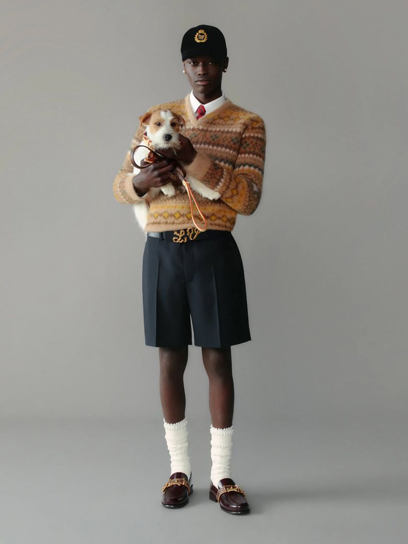 An adult male is holding a small dog in a full shot. The man is wearing a sweater and hat, while the dog is on a leash.