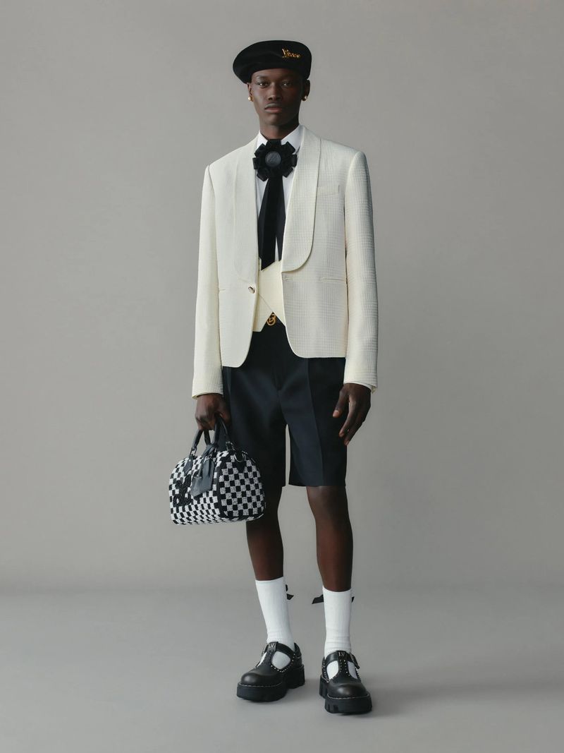 Male fashion model wears a cream blazer, black shorts, socks, and lug-soled T-strap shoes, holding a checkerboard satchel in a studio setting.