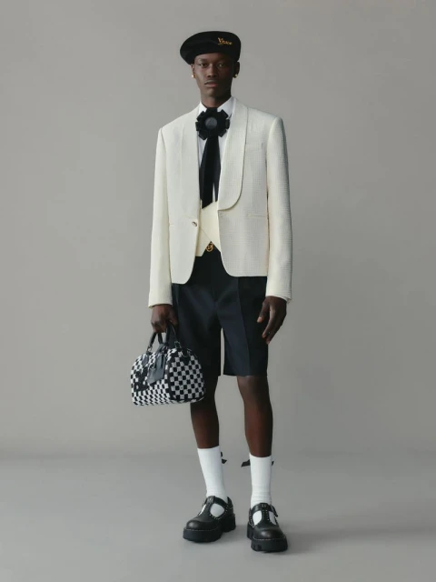Male fashion model wears a cream blazer, black shorts, socks, and lug-soled T-strap shoes, holding a checkerboard satchel in a studio setting.