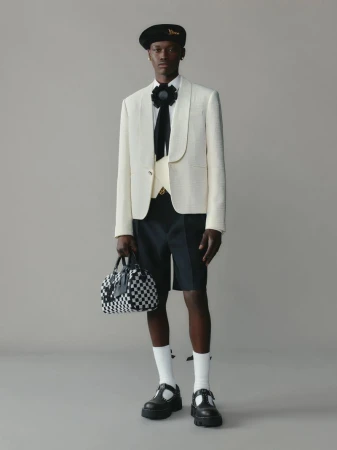 Male fashion model wears a cream blazer, black shorts, socks, and lug-soled T-strap shoes, holding a checkerboard satchel in a studio setting.