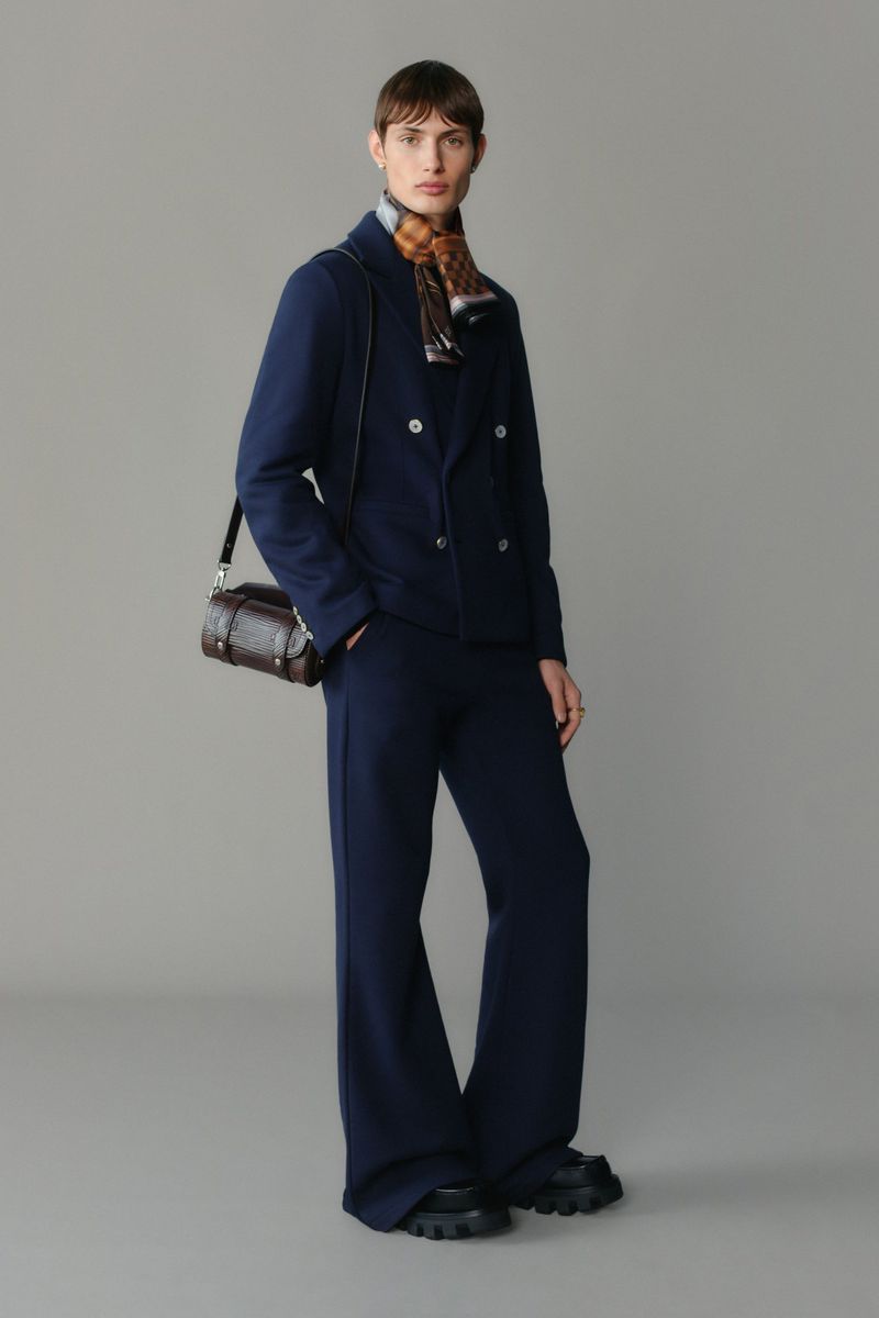 Full-length fashion shot of an adult woman in a navy suit and lug-soled shoes, carrying a brown leather handbag on a grey background.