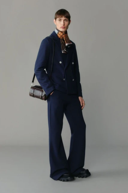 Full-length fashion shot of an adult woman in a navy suit and lug-soled shoes, carrying a brown leather handbag on a grey background.