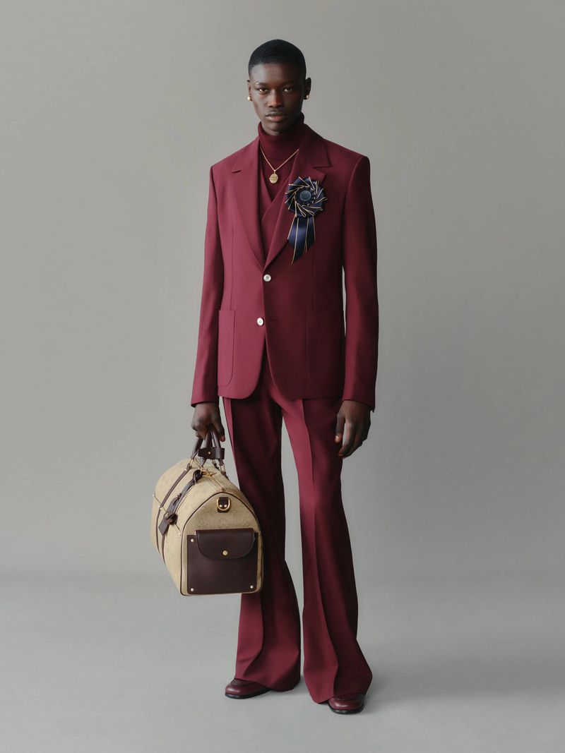 Male fashion model in a maroon suit with flared pants holding a tan duffel bag posing in a studio with a light grey background.