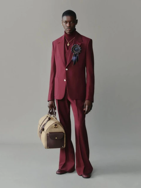 Male fashion model in a maroon suit with flared pants holding a tan duffel bag posing in a studio with a light grey background.