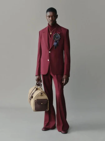 Male fashion model in a maroon suit with flared pants holding a tan duffel bag posing in a studio with a light grey background.