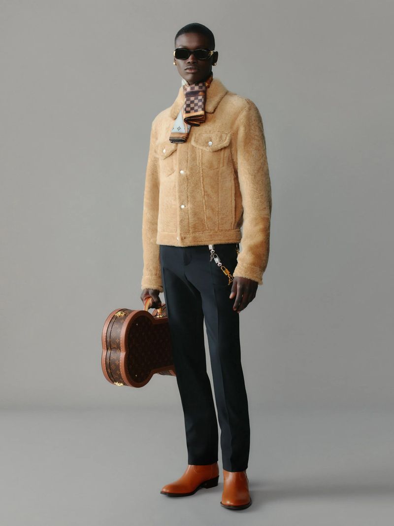Dark-skinned male model in a tan jacket, dark pants, and brown boots holding a brown Louis Vuitton guitar case. He also wears sunglasses and a scarf.