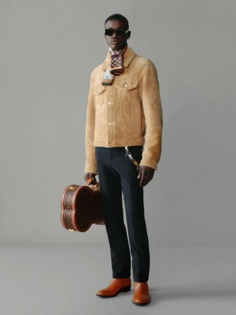 Dark-skinned male model in a tan jacket, dark pants, and brown boots holding a brown Louis Vuitton guitar case. He also wears sunglasses and a scarf.