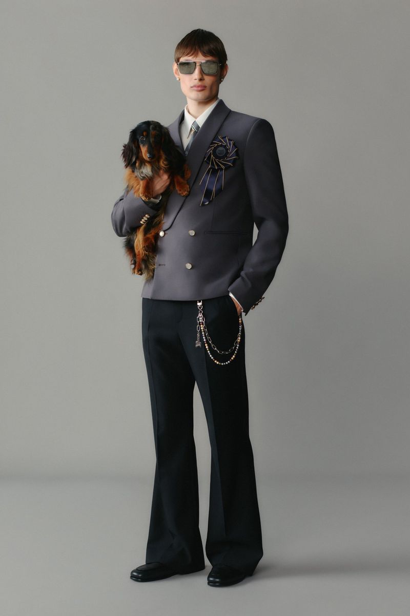 Stylish adult male in grey suit holds a long-haired dachshund dog, posing in a studio with black dress shoes and chain. Fashion editorial.