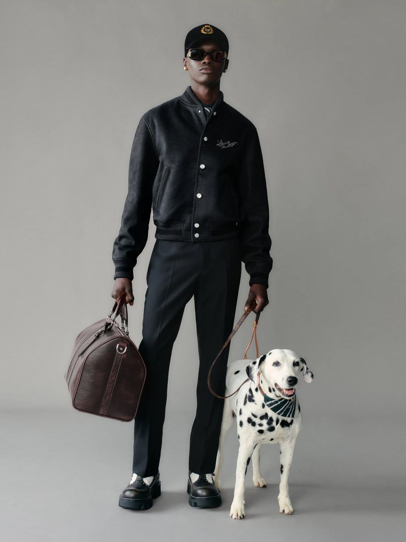 An adult Black man stands with a brown duffel bag and a Dalmatian dog in a studio, modelling a dark bomber jacket, trousers, and baseball cap.