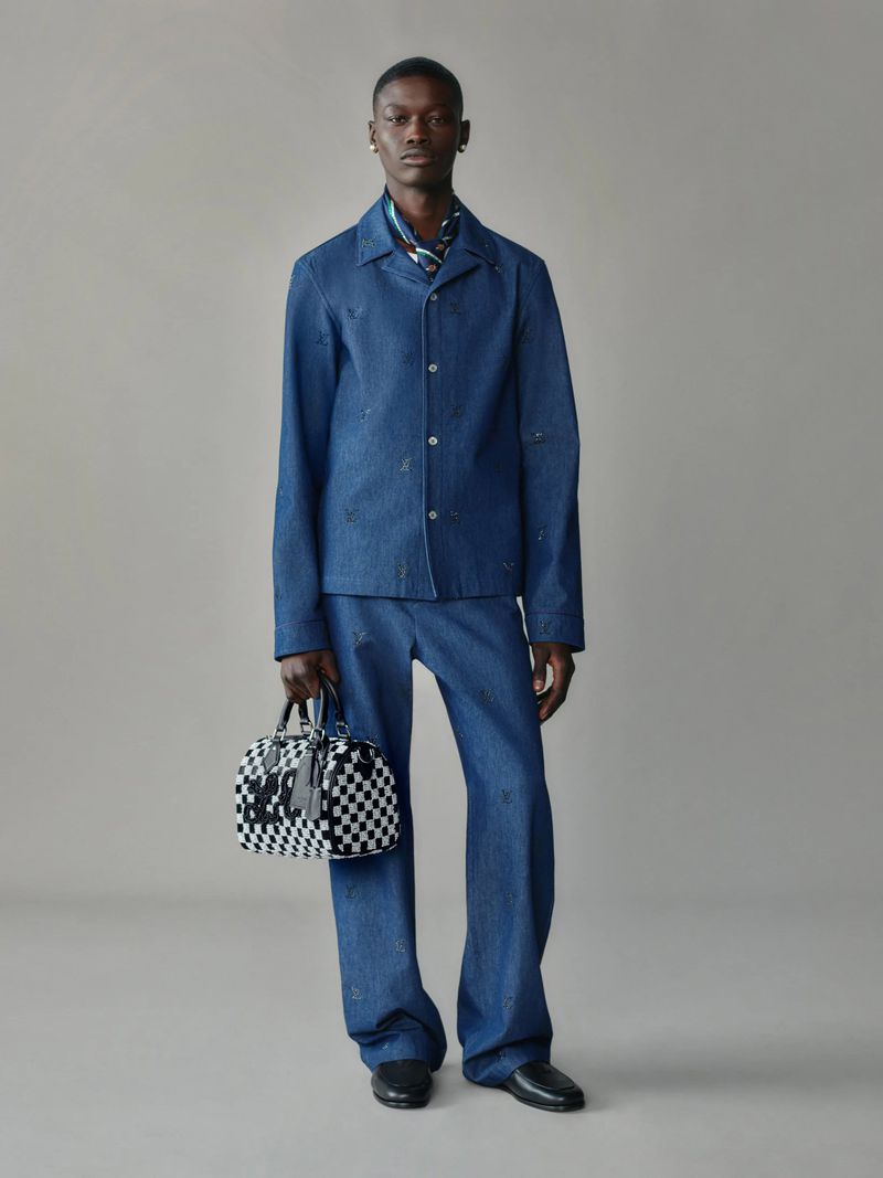 Full-length shot of an adult male fashion model in a denim outfit holding a checkered duffel bag against a plain background.